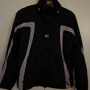 Svart skidjacka från Helly Hansen - Snygg svart vindjacka från Helly Hansen med vita reflex detaljer. Jackan har en hög krage och långa ärmar. Den har en sportig look perfekt för skidsäsongen