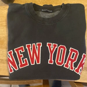 Svart sweatshirt från Brandy Melville - Snygg svart sweatshirt från Brandy Melville med fastsydda bokstäver texten 'New York' i rött och vitt på framsidan. Tröjan har långa ärmar och en rund halsringning. Perfekt för en avslappnad stil. Onesize/small