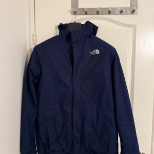 Blå vindjacka från The North Face - Säljer en blå vindjacka från The North Face med huva och dragkedja. Perfekt för blåsiga dagar och har en stilren design med logotyp på bröstet och ryggen. 