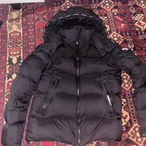 Svart dunjacka från Moncler - Säljer en snygg svart dunjacka från Moncler. Jackan har en pufferdesign med dragkedja och huva, perfekt för kyliga dagar. Den är långärmad och har en stilren look som passar till det mesta. Perfekt för dig som vill hålla värmen med stil!