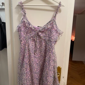 Lila blommig klänning med volangdetaljer - Charmig lila klänning med blommigt mönster och volangdetaljer. Den har smala axelband och en söt knytning framtill. Perfekt för en somrig look! Aldrig använd. 