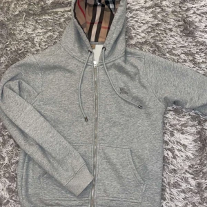 Grå hoodie från Burberry - Snygg grå hoodie från Burberry med klassiskt rutigt mönster i huvan. Tröjan har dragkedja och snörning vid huvan samt praktiska fickor framtill. Perfekt för en stilren och bekväm look.