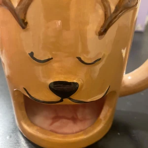 Brun mugg med renmotiv - Charmig brun mugg med ett sött renansikte och horn. Perfekt för varma drycker under vintern. Muggen har en unik design med en öppning i botten som ger den ett lekfullt uttryck.🤍