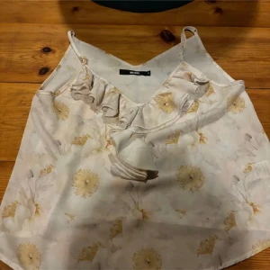 Blommig linne från Bik Bok - Säljer ett sött linne från Bik Bok med blommigt mönster i ljusa färger. Linnet har smala axelband och en volangdetalj framtill. Perfekt för varma sommardagar!
