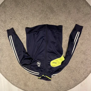 Mörkblå träningsjacka med gula detaljer - Snygg mörkblå träningsjacka från Adidas med gula detaljer och vita ränder längs ärmarna. Jackan har dragkedja och ett tryckt emblem på bröstet. Perfekt för sportiga aktiviteter. Säljs inte längre