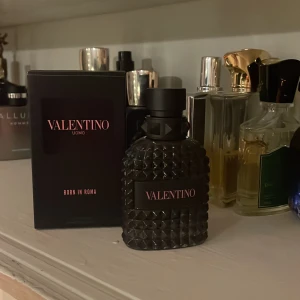 Valentino Uomo Born in Roma - Elegant parfymflaska från Valentino med en lyxig svart design och textur. Flaskan har en modern och stilren look med märkets logotyp i rosa. Perfekt för den som söker en sofistikerad doftupplevelse.