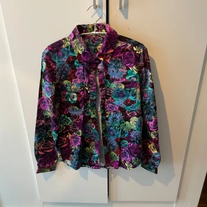 Blommig skjorta från Nelly Trend - Snygg blommig skjorta från Nelly Trend i storlek XS. Skjortan har ett färgglatt mönster med rosor i lila, rosa, grönt och gult. Den har långa ärmar och en knytning vid halsen för en extra detalj.