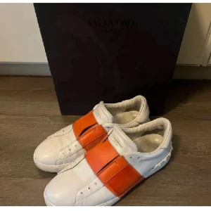 Valentino sneakers med orange detalj - Snygga vita sneakers från Valentino med en iögonfallande orange rem över ovansidan. Skorna har en klassisk design med rund tå och är tillverkade i skinn för en lyxig känsla.