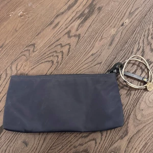 Svart kuvertväska från DKNY - Elegant svart kuvertväska från DKNY med en stilren design. Väskan har en avtagbar rem i guld och en diskret logotypdetalj. Perfekt för att bära det nödvändigaste med stil.