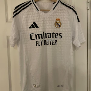 Vit Real Madrid fotbollströja - Snygg vit Real Madrid fotbollströja från Adidas med svart text och detaljer. Tröjan har korta ärmar och klubbens emblem på bröstet. Perfekt för alla fans som vill visa sitt stöd. De är den nysatte tröjan i storlek L men passar båda M bra 