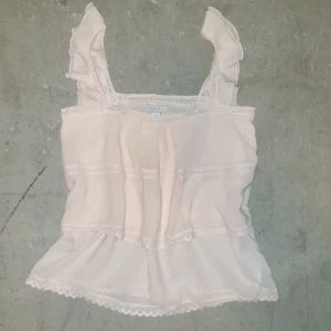 Puderrosa topp med spetsdetaljer - Säljer en söt rosa topp från Amisu med spetsdetaljer och volanger på axelbanden. Toppen har en luftig passform och är perfekt för varma dagar. Den är tillverkad i ett lätt och bekvämt material.