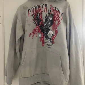 Grå hoodie från Black Squad - Säljer en grå hoodie från Black Squad med tryck på baksidan. Hoodien har en stor örn och texten 'Pioneer Souls' i rött och svart. Den är i oversized passform och har en klassisk känguruficka framtill.