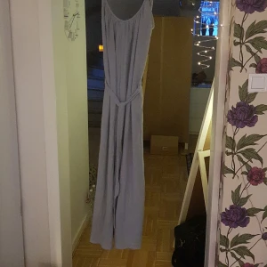 Ljusgråblå byxklänning från H&M - Elegant blågrå byxklänning från H&M med ett knytband i midjan. Klänningen har en v-ringad design och är ärmlös, vilket ger en stilren och luftig look. Perfekt för sommarens festligheter.
