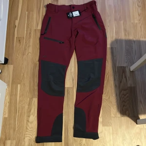Röda byxor med svarta detaljer - Snygga röda byxor från Pinewood med svarta förstärkningar på knän och bak. De har flera praktiska dragkedjefickor och är tillverkade i ett snabbtorkande material. Perfekta för utomhusaktiviteter.