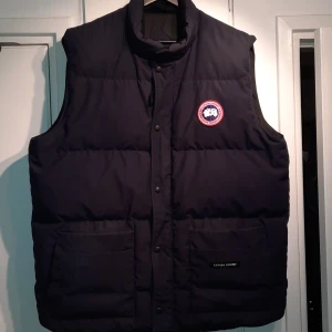 Marinblå dunväst från Canada Goose - Säljer en svart dunväst från Canada Goose med broderad logotyp på bröstet. Västen har knappar och två stora fickor framtill. Perfekt för kyliga dagar. (ÄKTA) inget snack om saken. Pris kan diskuteras! Förhoppningsvis så passar västen även dig som har XL