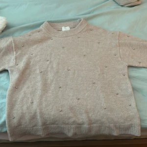 Beige/rosa tröja med pärlor från H&M - Säljer en stilren beige tröja från H&M dekorerad med små pärlor. Tröjan har en rund hals och korta ärmar, perfekt för en elegant vardagslook. Passar bra till både jeans och kjol.