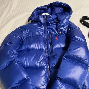 Blå dunjacka från Polo Ralph Lauren - Snygg blå dunjacka från Polo Ralph Lauren med dragkedja och huva. Perfekt för kyliga dagar med sin vadderade design. Jackan har en liten röd logotyp på bröstet och praktiska fickor.