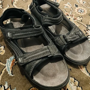 Svarta sandaler med kardborreband - Säljer ett par svarta sandaler från iN2 med justerbara kardborreband. De har en robust sula och är perfekta för sommarens äventyr. Sandalerna är bekväma och lätta att ta på och av.