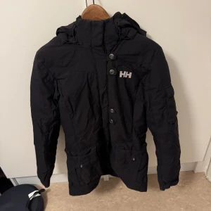 Svart jacka från Helly Hansen - Säljer en svart jacka från Helly Hansen med huva och knappar. Perfekt för kyligare dagar med sin praktiska design och flera fickor. Helly Hansen-logga på bröstet ger en sportig touch.