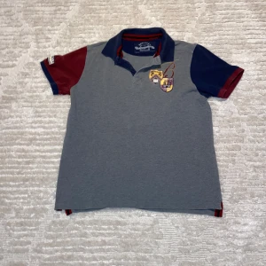 Grå pikétröja från Berkeley - Snygg grå pikétröja från Berkeley med broderade emblem på bröstet och ärmen. Tröjan har en klassisk krage med röd och blå randig detalj. Ärmarna är korta och i kontrasterande röd och blå färg.