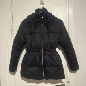 Svart pufferjacka med huva - Snygg svart pufferjacka med dragkedja och justerbar midja. Jackan har en praktisk huva och långa ärmar, perfekt för kyliga dagar. Stilren design som passar till det mesta.