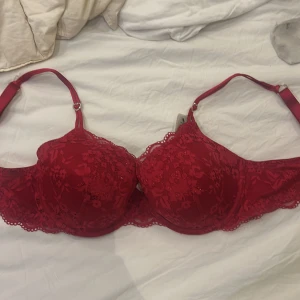 Röd spets-bh med hjärtformad detalj - Snygg röd bh i spets med en glittrig hjärtformad detalj på axelbandet. Den har ingen push upp och justerbara axelband för bästa passform. Perfekt för en romantisk look. Aldrig använd, prislapp kvar på.
