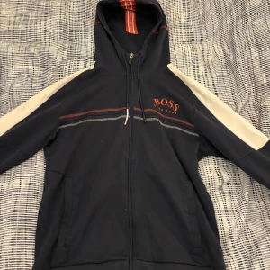 Mörkblå hoodie från BOSS - Snygg mörkblå hoodie från BOSS med dragkedja och huva. Den har vita detaljer på ärmarna och ett rött BOSS-tryck på bröstet. Perfekt för en avslappnad stil. storlek M men passar S