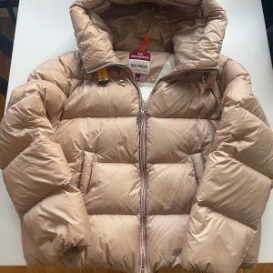 Beige dunjacka från Parajumpers - Säljer en snygg beige dunjacka från Parajumpers med huva och dragkedja. Jackan har en klassisk pufferdesign och är perfekt för kyliga dagar. Märkesdetaljer på ärmen ger en exklusiv touch.