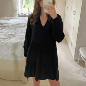 Svart klänning med v-ringning - Snygg svart klänning med v-ringad hals och långa ärmar. Perfekt för en stilren look. Klänningen har en avslappnad passform och faller fint.