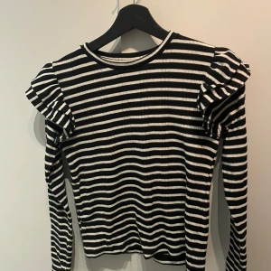 Randig tröja från Zara - Säljer en svartvit randig tröja från Zara med volangdetaljer på axlarna. Tröjan har långa ärmar och en rund halsringning. Perfekt för en stilren look!