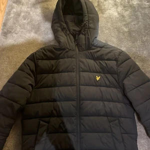 Mörk blå pufferjacka från Lyle & Scott - Snygg mörk blå pufferjacka från Lyle & Scott med huva och dragkedja. Jackan har ett broderat emblem på bröstet och är perfekt för kyligare dagar. Den är långärmad och har en stilren design. Köpte för 980 kr