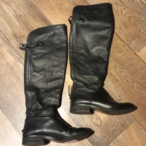 Svarta skinnstövlar - Snygga svarta höga stövlar i skinn med dragkedja och spänne på sidan. Perfekta för höst- och vinterbruk med en stilren design som passar till många olika outfits.