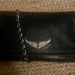 Zadig Voltaire rock väska  - Snygg svart axelväska i skinn med en cool kedja som axelrem. Båda banden finns. Väskan har en dragkedja på baksidan och en dekorativ detalj i form av vingar på framsidan. Perfekt för att ge din outfit en edgy touch.🖤🖤🖤