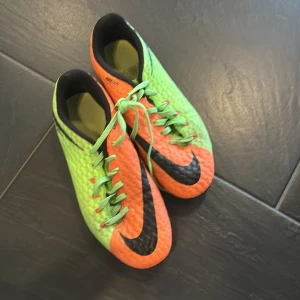Grön och orange fotbollsskor från Nike - Säljer ett par iögonfallande fotbollsskor från Nike i grön och orange med svarta detaljer. Skorna har snörning och dobbar för bra grepp på planen. Perfekta för fotbollsspelare som vill sticka ut!