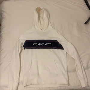 Vit hoodie från GANT - Säljer en stilren vit hoodie från GANT med en stor logga på bröstet i marinblått och rött. Hoodien har en klassisk känguruficka och en bekväm huva. Perfekt för en avslappnad stil.