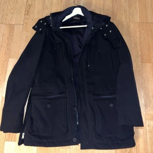 Jacka från Oscar Jacobson - Säljer en stilren svart parkajacka från Oscar Jacobson. Jackan har långa ärmar och praktiska fickor med knappar framtill. Den har en dragkedja och knappar för extra skydd mot väder och vind. Perfekt för kyligare dagar.