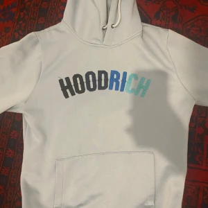 Ljusgrå hoodie och byxor från Hoodrich - Säljer ett set med ljusgrå hoodie och matchande byxor från Hoodrich. Hoodien har en stor ficka framtill och texten 'HOODRICH' i svart och blått på bröstet. Byxorna har samma text på ena benet. 