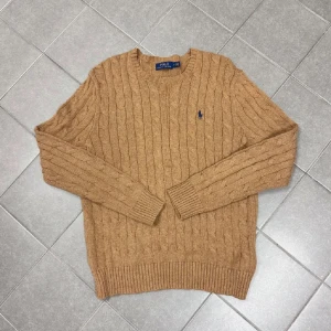 Beige stickad tröja från Polo Ralph Lauren - Säljer en snygg beige stickad tröja från Polo Ralph Lauren med kabelstickat mönster. Tröjan har långa ärmar och en liten broderad logga på bröstet. Perfekt för kyligare dagar!
