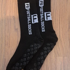 FS Gripsocks - Säljer ett par svarta fotbollsstrumpor från FS med vit text. Strumporna har greppytor under fötterna för bättre fäste. Perfekta för fotbollsträning eller match.