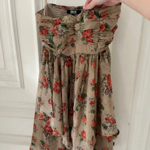 Blommig klänning från ASOS - Charmig blommig klänning från ASOS i beige med röda och gröna blommor. Klänningen har en smockad överdel och är axelbandslös, vilket ger en romantisk känsla. Perfekt för sommarens varma dagar. 💕