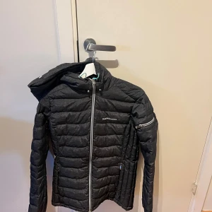 Svart dunjacka från Peak Performance Dam - Säljer en svart dunjacka från Peak Performance med dragkedja och huva. Jackan har en quiltad design och är perfekt för kyligare väder. Den har långa ärmar och en stilren look. Passar bra för både stad och natur. Jackan har användts 2 gånger och är 5 år gammal