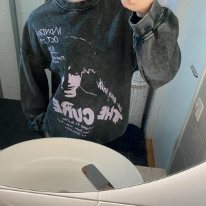 Svart sweatshirt med The Cure-tryck - Cool svart sweatshirt med stort The Cure-tryck i vitt och lila på framsidan. Tröjan har en avslappnad passform och långa ärmar, perfekt för en casual look. Den har en vintage tvättad stil som ger en unik känsla.
