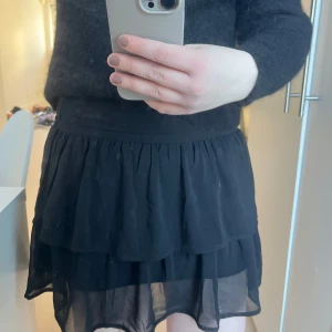 Svart kortkjol med volang - Snygg svart kortkjol med volangdetaljer. Perfekt för att skapa en chic look. Kjolen har en luftig design och är lätt att matcha med olika toppar. 