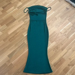 Grön långklänning med smala axelband - Elegant grön långklänning med smala axelband och en figurnära passform. Klänningen har en stilren design som framhäver silhuetten och är perfekt för festliga tillfällen.