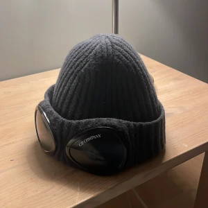  C.P. Company mössa - Snygg grå ribbad mössa från C.P. Company med unika glasögondetaljer framtill. Perfekt för att hålla värmen och samtidigt se stilren ut. Den är i bra skick och passar perfekt till allt