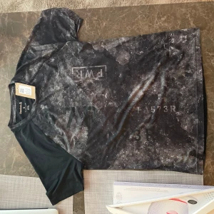 Svart och grå t-shirt från Reebok - Snygg svart och grå t-shirt från Reebok med ett unikt mönster. Perfekt för träning eller en avslappnad dag. T-shirten har korta ärmar och är tillverkad i ett lätt och bekvämt material.