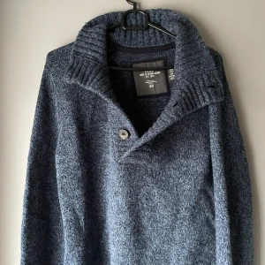 Mörkblå stickad tröja från H&M - Säljer en mörkblå stickad tröja från H&M med en stilren design. Tröjan har en hög krage med knappdetalj och långa ärmar. Perfekt för kyligare dagar.