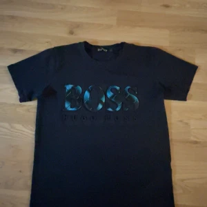 Svart t-shirt från Hugo Boss - Snygg svart t-shirt från Hugo Boss med ett stort, glansigt BOSS-tryck i blått och svart på framsidan. Perfekt för en stilren look.