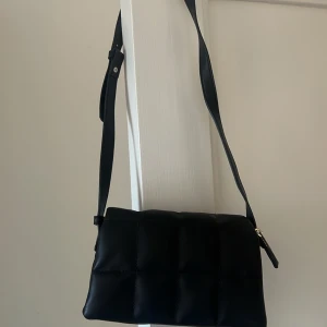 Svart quiltad axelväska - Snygg svart axelväska med quiltad design. Väskan har en justerbar axelrem och en stilren look som passar perfekt till olika outfits. Perfekt för att bära med sig det nödvändigaste med stil.