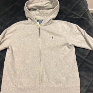 Grå hoodie från Polo Ralph Lauren | L 14-16 år - Säljer en stilren grå hoodie från Polo Ralph Lauren med dragkedja och huva. Tröjan har en liten broderad logga på bröstet och är perfekt för en avslappnad look. Passar både till vardags och chill.